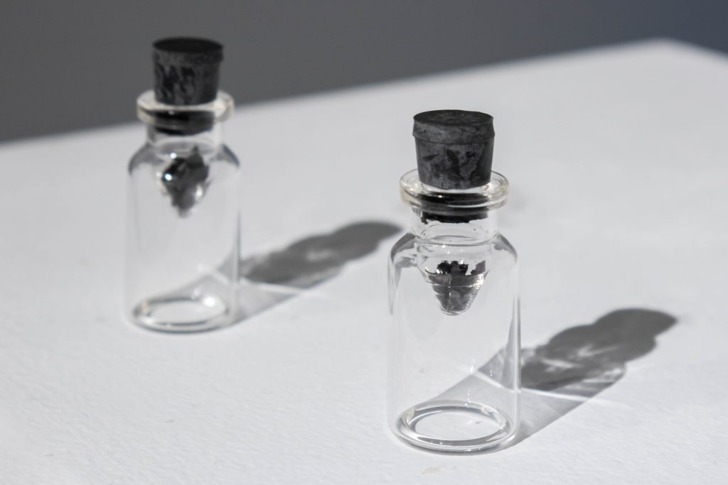 A closeup of two small glass jars with black rubber stoppers, about 3 inches in height. The jars, instead of their whole interior being the chamber that can hold material, have smaller, conical chambers within. The conical chambers hold blackened flecks of dried blood, and the spotlight above is refracted by the glass and winks like a star across the white pedestal on which the jars sit.