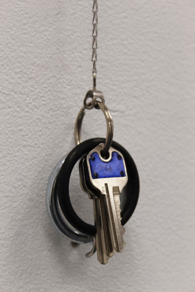 A retractable key ring is mounted on a white wall. The silver disc that houses the chain and mechanism is at the top, while the key ring with its cluster of objects is below, held on a hook. The chain between them is taut, pulling. The objects on the key ring include a key with a blue head, a rubber strap-on O-ring, and a steel cock ring.