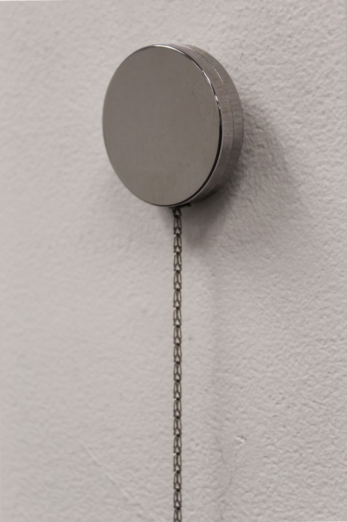 A retractable key ring is mounted on a white wall. The silver disc that houses the chain and mechanism is at the top, while the key ring with its cluster of objects is below, held on a hook. The chain between them is taut, pulling. The objects on the key ring include a key with a blue head, a rubber strap-on O-ring, and a steel cock ring.