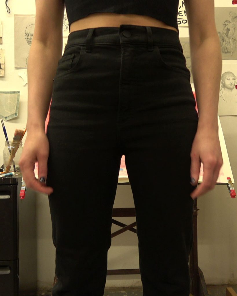 A still from a video. The artist stands in their studio, wearing black pants and a black cropped tank top. We see from their mid-torso down to their knees. Their hands hang limply at their sides.