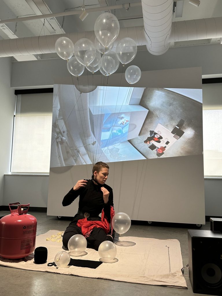 A photo from a performance. The framing is wide. The artist kneels on a bed sheet. Behind them is a projection of the room—but it's tilted and shows the projection, so it therefore spirals in, smaller and smaller, in a mise en abyme. In front of the artist on the bed sheet is a helium tank, clear latex balloons, and black thread. The artist is dressed all in black, save for a red silk scarf thrown over their left shoulder. They are in the process of filling up the balloons with helium and tying them to a needle-filled necklace around their neck.