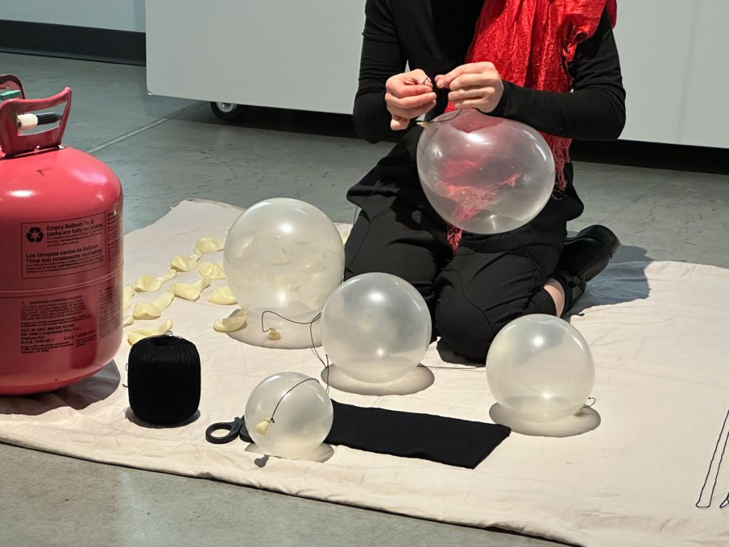 A photo from a performance. The artist kneels on a bed sheet. In a closeup of their hands, they are in the process of tying a black string to a clear latex balloon. The balloon bulges, and less-full balloons hover before the artist. On the bed sheet are rows of black strings, unfilled balloons, and a red helium tank. The artist wears all black, except for a red silk scarf on their left shoulder.