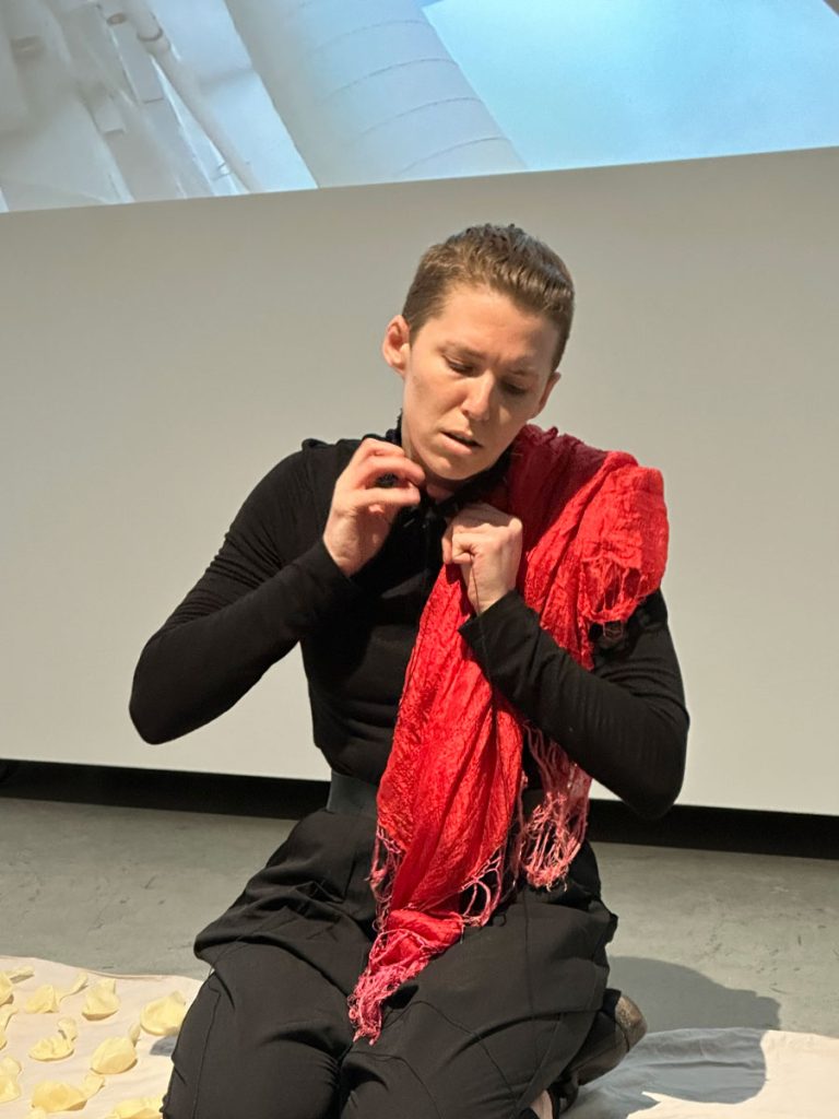 A photo from a performance. The artist kneels on a bed sheet. In a closeup, the artist is tying a black thread necklace to their neck. The necklace is full of needles. The artist looks distraught, or not fully aware.