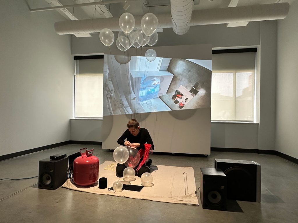 A photo from a performance. In a wide shot, the artist kneels on a bed sheet. A spray of balloons floats up from their neck, where they are tied to a necklace full of needles.