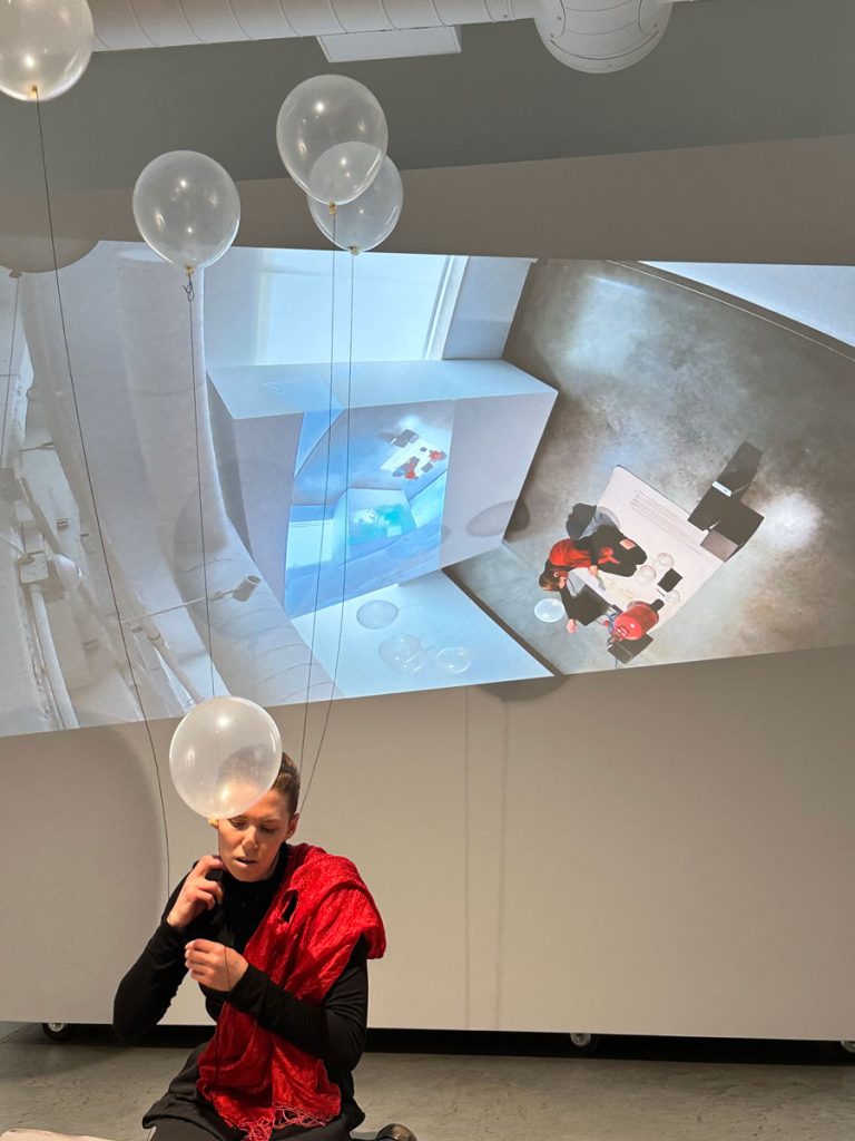 A photo from a performance. The artist kneels on a bed sheet. Behind them is a projection of the room—but it's tilted and shows the projection, so it therefore spirals in, smaller and smaller, in a mise en abyme. The artist is dressed all in black, save for a red silk scarf thrown over their left shoulder. They are in the process of filling up the balloons with helium and tying them to a needle-filled necklace around their neck. One of the clear latex balloons floats up in front of their face, partially obscuring it.