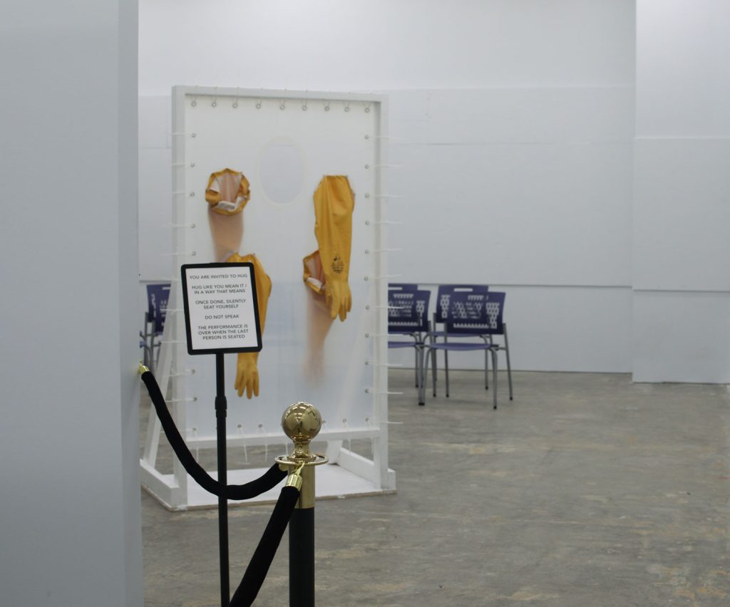An establishing image of a sculpture necessary for the performance, the hug machine. Stanchions with black rope lead to a sign that says "You are invited to hug. Hug like you mean it / in a way that means. Once done, silently seat yourself. Do not speak. The performance is over when the last person is seated." Behind the sign, the hug machine waits: a freestanding door frame made of white rubberized wood. A grommeted silicone sheet is suspended in the frame with zipties. Two long yellow pairs of rubber gloves penetrate the sheet, and there is a hole approximately face-height about 50 inches high. Two people could hug one another through the barrier using the gloves, and the hole would let them be face-to-face. Behind the hug machine sits rows of dark blue chairs in an otherwise empty white room with concrete floors.