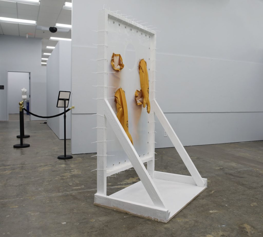 A different view of the hug machine: a freestanding door frame made of white rubberized wood. A grommeted silicone sheet is suspended in the frame with zipties. Two long yellow pairs of rubber gloves penetrate the sheet, and there is a hole approximately face-height about 50 inches high. Two people could hug one another through the barrier using the gloves, and the hole would let them be face-to-face.