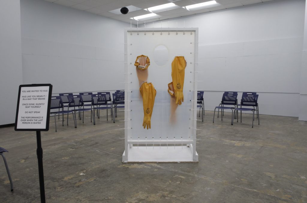 A different view of the hug machine: a freestanding door frame made of white rubberized wood. A grommeted silicone sheet is suspended in the frame with zipties. Two long yellow pairs of rubber gloves penetrate the sheet, and there is a hole approximately face-height about 50 inches high. Two people could hug one another through the barrier using the gloves, and the hole would let them be face-to-face. Behind the hug machine sits rows of dark blue chairs in an otherwise empty white room with concrete floors.